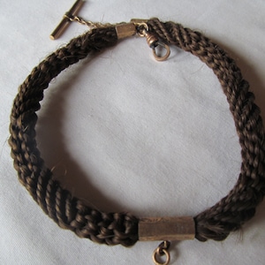 May include: A brown hair chain with a gold clasp and a gold T-bar. The chain is braided and has a gold bar in the center.