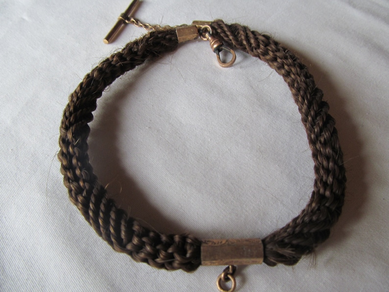 May include: A brown hair chain with a gold clasp and a gold T-bar. The chain is braided and has a gold bar in the center.