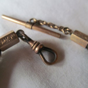 May include: A gold pocket watch chain with a brown leather tassel. The chain has a decorative clasp and a swivel hook.