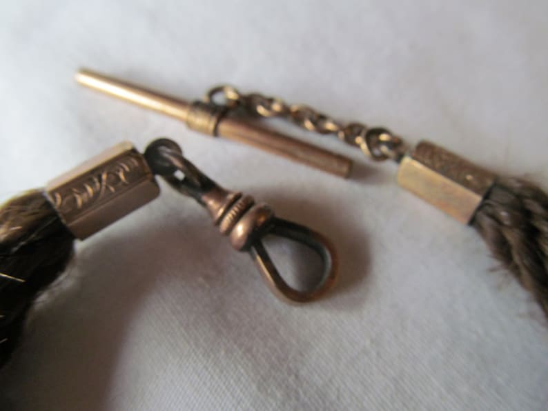 May include: A gold pocket watch chain with a brown leather tassel. The chain has a decorative clasp and a swivel hook.