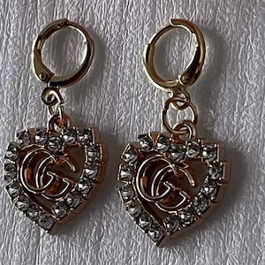 May include: A pair of gold-tone earrings with a heart-shaped design. The hearts are adorned with clear rhinestones and have a double 'G' logo in the center.