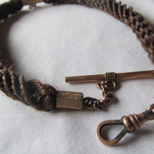 May include: A braided hair watch chain with a gold clasp and a swivel hook. The chain is made of brown hair and is about 12 inches long.