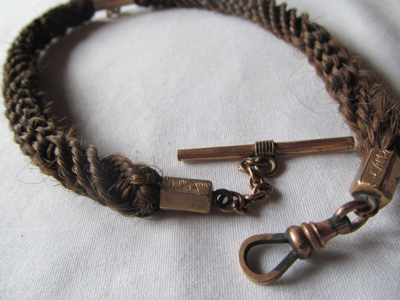 May include: A braided hair watch chain with a gold clasp and a swivel hook. The chain is made of brown hair and is about 12 inches long.