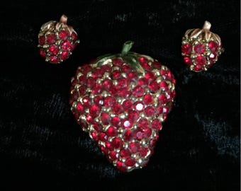 May include: A strawberry-themed jewelry set featuring a brooch and matching earrings. The brooch and earrings are shaped like strawberries, covered in red rhinestones, and have gold-toned metal accents. The set is displayed on a black background.