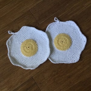 May include: Two handmade crochet potholders shaped like fried eggs. The potholders have a white background with a yellow center, and a small loop for hanging. The potholders are on a wooden surface.