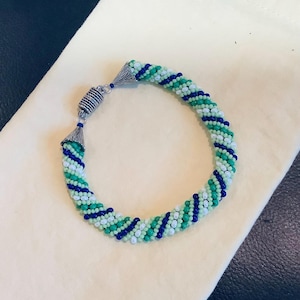 May include: A beaded bracelet with a spiral design. The bracelet features alternating rows of green, white, and navy blue beads. The clasp is silver-toned with a magnetic closure. The bracelet is resting on a cream-colored surface.