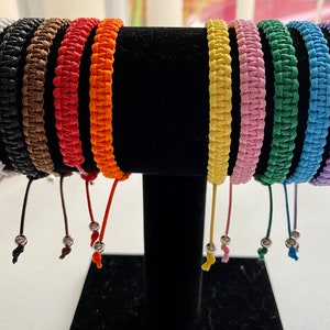 May include: A set of 10 colourful braided bracelets with silver beads. The bracelets are made of woven string and come in a variety of colours, including white, black, brown, red, orange, yellow, pink, green, blue, and purple.