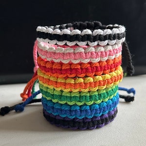 May include: A stack of colourful friendship bracelets made with woven string. The bracelets are in a rainbow pattern with black and white stripes at the top and bottom.