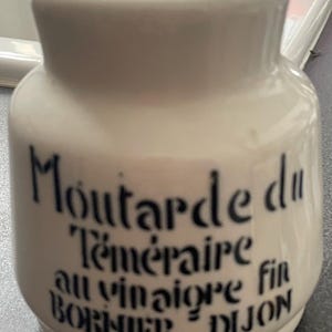 Puede incluir: Bote de mostaza de cerámica blanquecina con un borde ligeramente ensanchado. El bote tiene texto negro que dice "Moutarde du Téméraire au vinaigre fin BORNIER DIJON". Forma cilíndrica con base redondeada.