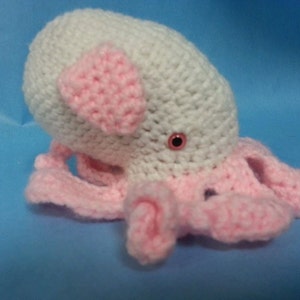Puede incluir: Un pulpo de ganchillo hecho a mano en blanco y rosa. El pulpo tiene una cabeza blanca con detalles rosas en forma de orejas y tentáculos rosas. Los ojos son pequeños y rojos. El juguete está sobre una superficie azul.