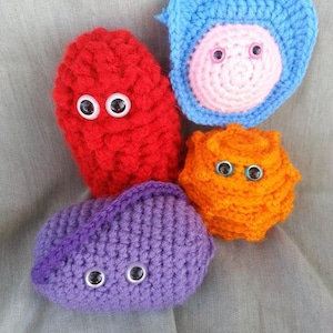 May include: Four crocheted plush toys with googly eyes. The toys are shaped like a ghost, a square, a spiky ball, and a bean. The ghost is red, the square is blue with a pink center, the spiky ball is orange, and the bean is purple.