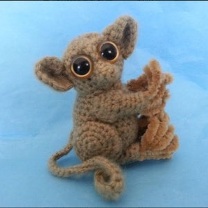 Puede incluir: Una muñeca tarsio de ganchillo hecha a mano en tonos marrones y beige. La muñeca tiene ojos grandes y expresivos, una cola larga y está posada con los brazos extendidos. El fondo es de color azul claro liso.