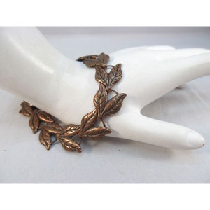 May include: A copper-toned bracelet featuring a leaf design. The bracelet is composed of multiple leaf-shaped links connected by small rings. The leaves have detailed veining and a warm, metallic finish. The bracelet is displayed on a white surface.