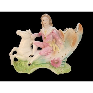 May include: A porcelain figurine of a person riding a white horse. The figurine is painted in pink, green, and gold. The horse is standing on a green base with a pink flower.