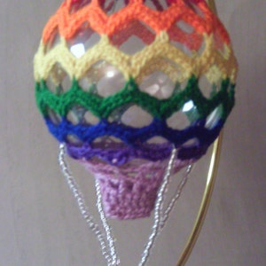 May include: A crocheted hot air balloon ornament with a rainbow pattern and a small basket. The ornament is hanging from a gold stand.