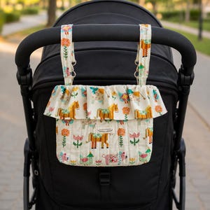 May include: A cream-colored quilted stroller organizer bag featuring a colorful pattern of horses, pigs, chickens, and flowers. The bag has a ruffled top edge and two fabric straps with metal rings for attachment. A small white label reads Dream Living BC.