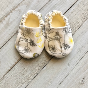 May include: A pair of white baby shoes with a yellow and gray floral print featuring bees and mason jars.
