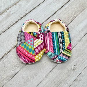 May include: A pair of baby booties with a colorful geometric pattern. The booties feature a patchwork design with pink, teal, yellow, and gray shapes. The interior is a soft, cream-colored fabric, and the edges are elasticized.