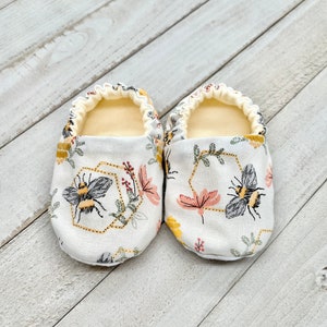 May include: A pair of white baby booties with a bee and honeycomb pattern. The booties have a yellow, black, and pink floral design.