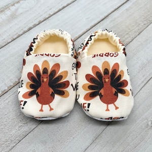 May include: A pair of baby booties with a white background and a brown turkey print. The booties have the text "Gobble 'til we wobble!" printed on them.