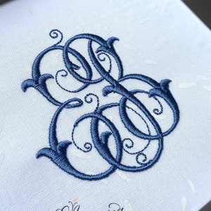 May include: White square fabric with a large, ornate navy blue monogram. The monogram features intertwined letters with decorative flourishes. The fabric has a subtle pattern and the text "A. namorel by ANAM GLAM INC" is at the bottom.
