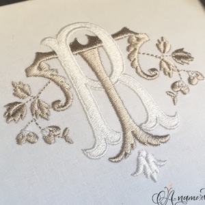 Może przedstawiać: Haftowany monogram litery "R" w kolorze białym i beżowym, z kwiatowymi zdobieniami. Monogram znajduje się na białym tle.
