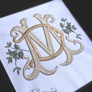 Könnte beinhalten: Ein Monogramm-Stickmuster mit den Buchstaben "M" und "D" in einer dekorativen Schriftart mit floralen Verzierungen. Die Buchstaben sind beigefarben und die Blumen sind in Grüntönen gehalten.