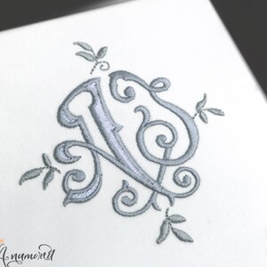 Peut inclure: Un monogramme brodé argenté de la lettre "N" avec des accents floraux décoratifs.