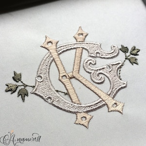 May include: A monogram embroidery design featuring the letters "K" and "G" in a decorative, intertwined style. The letters are embroidered in a light beige color on a white fabric background. The design is accented with small green floral sprigs.