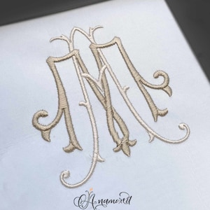 May include: A monogram with the letter "M" repeated three times in a decorative script font. The letters are embroidered in a light brown color on a white background.