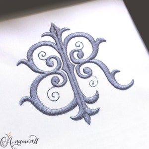 Może przedstawiać: Haftowany monogram w kolorze srebrnym z literą "R" w ozdobnym kroju pisma. Monogram znajduje się na białym tle.