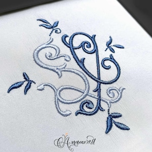 May include: Blue and silver embroidery design of a stylized letter "S" with floral accents and dragonfly motifs. The design is on a white fabric background. The text "Anamored" is written in cursive below the design.