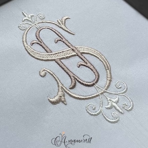 Könnte beinhalten: Ein weißer Stoff mit einem beige und weißen gestickten Monogramm des Buchstabens "S". Das Monogramm ist in einer Schriftart mit kunstvollen Verzierungen gehalten. Der Text "Anamored by ANAM GLAM INC" befindet sich am unteren Rand des Bildes.