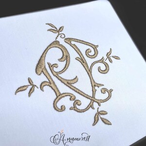 May include: A gold embroidered monogram of the letters "R" and "V" intertwined with floral designs. The monogram is on a white background.