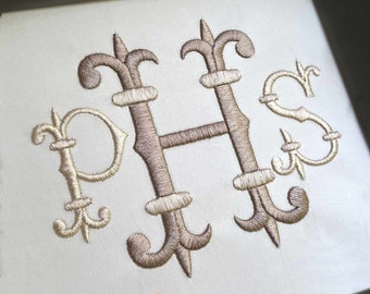 Puede incluir: Una superficie de tela blanca con un monograma grande bordado a máquina de las letras P, H y S en hilo de bronce metálico. Las letras están decoradas con adornos inspirados en la flor de lis. El texto Anamorei aparece debajo.