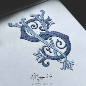 May include: Embroidered monogram in shades of blue, featuring the intertwined letters "S" and "V" with ornate floral accents. The design is on a light blue fabric, with the text "by ANAMOREL" and "by ANAM GLAM INC" at the bottom.