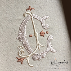 May include: A white embroidered monogram of the letter "C" with a decorative flourish. The embroidery is done in a traditional style with a light brown thread. The monogram is on a beige linen fabric.