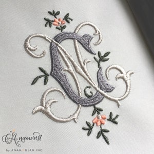 May include: A monogrammed embroidery design featuring the letter "C" in a gray color with a white outline. The letter is surrounded by a decorative floral design with green leaves and pink flowers.