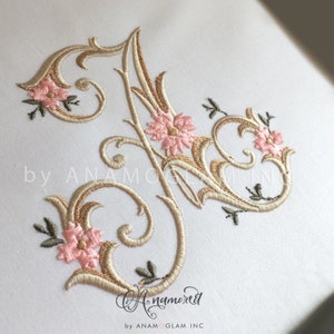 May include: Embroidered letter "J" in a gold and cream color with pink floral accents. The letter is in a decorative script font and is surrounded by intricate scrollwork. The background is white.