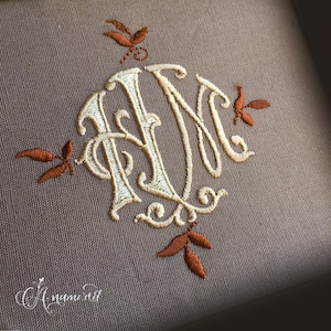 May include: A monogram with the letters "RM" in a white thread, surrounded by a circle of brown thread. The monogram is decorated with four small brown leaf designs.