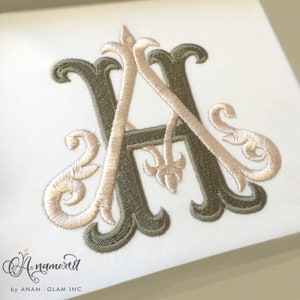 May include: Embroidered monogram featuring the letters "A" and "A" in a decorative, intertwined design. The letters are in a light tan color, outlined with a darker olive green. The design includes ornate flourishes and a fleur-de-lis element. The text "Anamored" is in the bottom left corner.