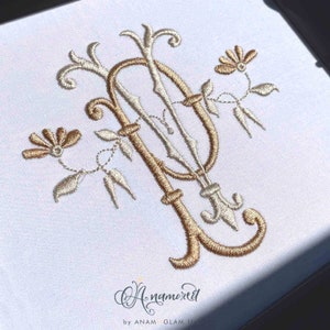 May include: A gold embroidered monogram of the letter "P" with floral embellishments. The monogram is surrounded by delicate white embroidery on a white fabric background.