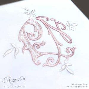 Puede incluir: Primer plano de una superficie blanca con un monograma bordado en rosa con las letras "A" y "N" entrelazadas con detalles florales. El diseño está rodeado de delicados motivos de hojas. El texto "A.namored" está en la parte inferior.