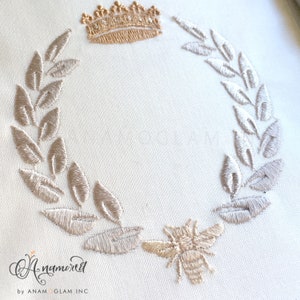 May include: A gold crown sits atop a wreath of leaves, with a small gold bee at the bottom of the wreath. The design is embroidered on a white fabric.