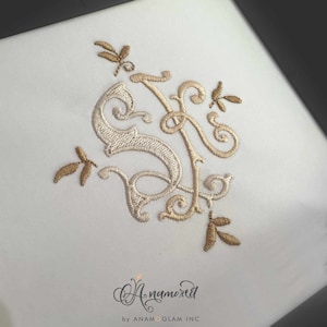 May include: A white fabric with a gold and cream embroidered monogram of the letters "S" and "F" with a decorative floral design. The monogram is surrounded by four small embroidered dragonfly designs.