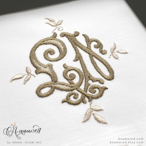 May include: A white fabric featuring a centered, intricate monogram embroidery in a metallic gold thread. The design consists of swirling, ornate letters surrounded by small leaf-like accents. The text Anamored by ANAMOGLAM INC appears below.