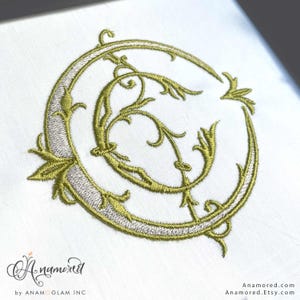 May include: Embroidered monogram in gold and silver thread on white fabric. The letter "C" is stylized with ornate floral and leaf details. The design is framed by a circular border. The Etsy shop name "Anamored" is in the bottom left corner.