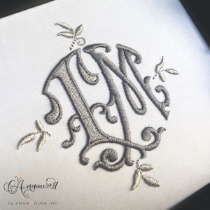 Puede incluir: Un monograma bordado en plata con las letras "LM" en una fuente de letra cursiva decorativa. El monograma está rodeado por cuatro pequeños diseños florales.