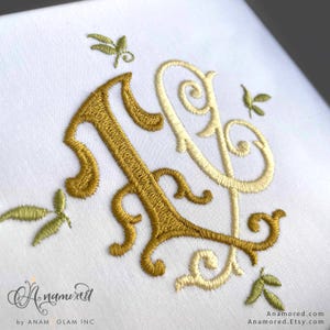May include: White fabric with embroidered gold and cream-colored initials, possibly a monogram. The design includes decorative flourishes and small green leaf accents. The embroidery is detailed and elegant, suggesting a personalized item.