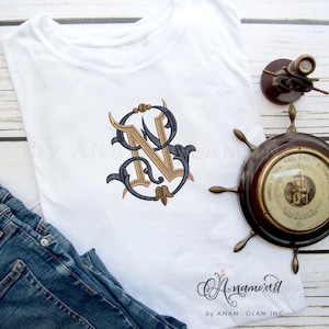 Puede incluir: Una camiseta blanca con un monograma bordado en oro y azul de la letra "N" en la parte delantera. La camiseta está sobre una superficie de madera con un par de pantalones vaqueros azules y una brújula vintage.